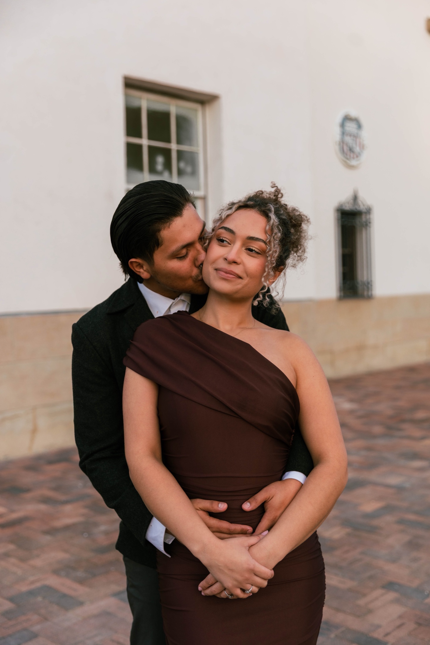 He kisses her cheek from behind in golden evening light